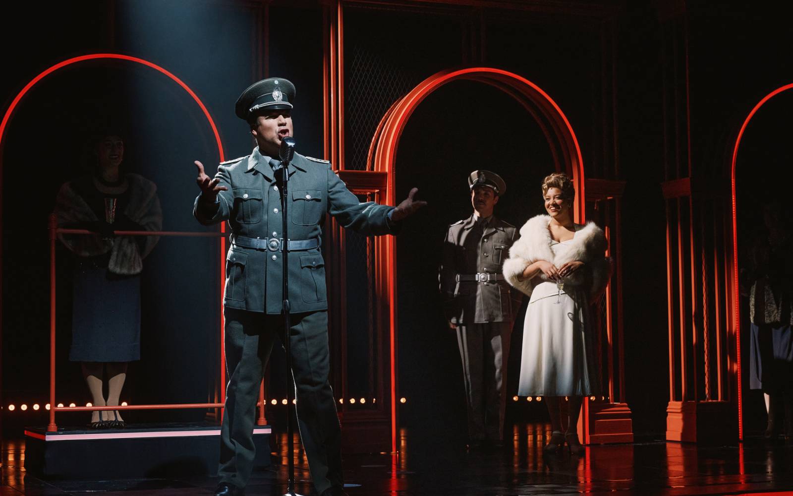 Caesar Samayoa as Perón speaks into a microphone, gesturing dramatically while Caleb Marshall-Villarreal, and Shereen Pimentel (Eva) look on.