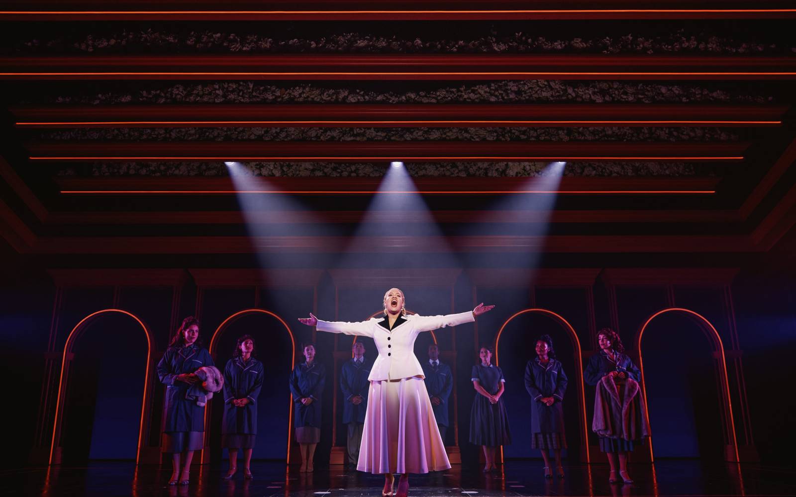 Shereen Pimentel as Eva standing in the spotlight with her arms outstretched while cast members look on.