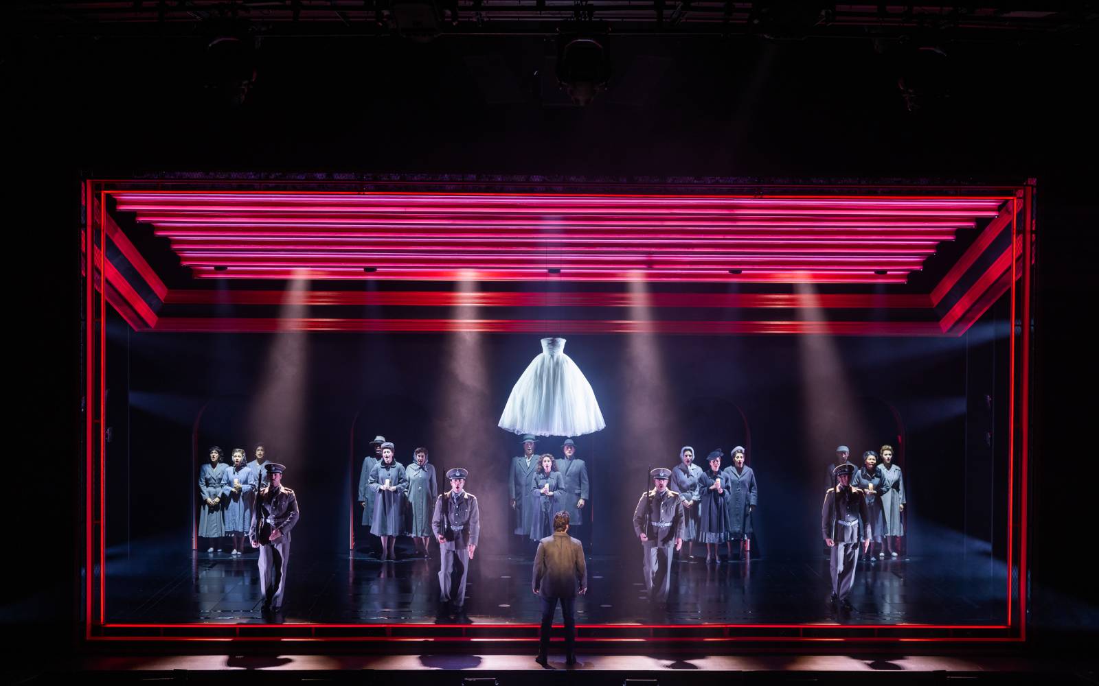 The company of Evita sings on stage, standing in small groups. They are framed by red lights and a white dress hangs overhead.
