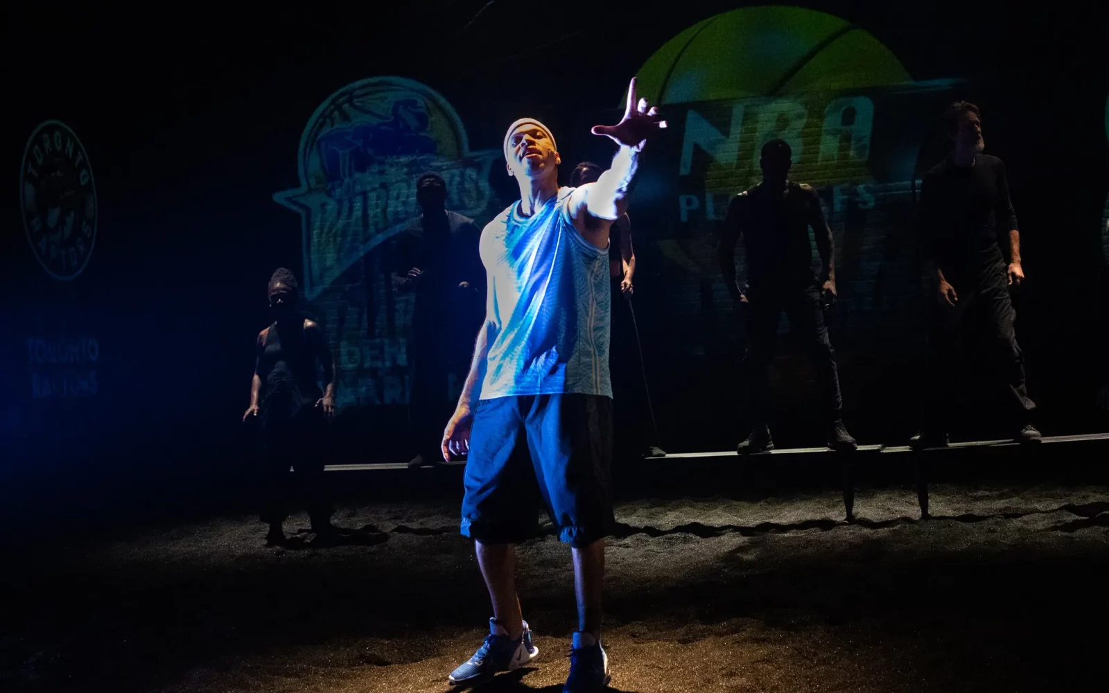 23Basketball-03-bkjp-superJumbo A man in a blue basketball jersey and shorts stands center stage in a spotlight, with a few figures in shadow in the background.