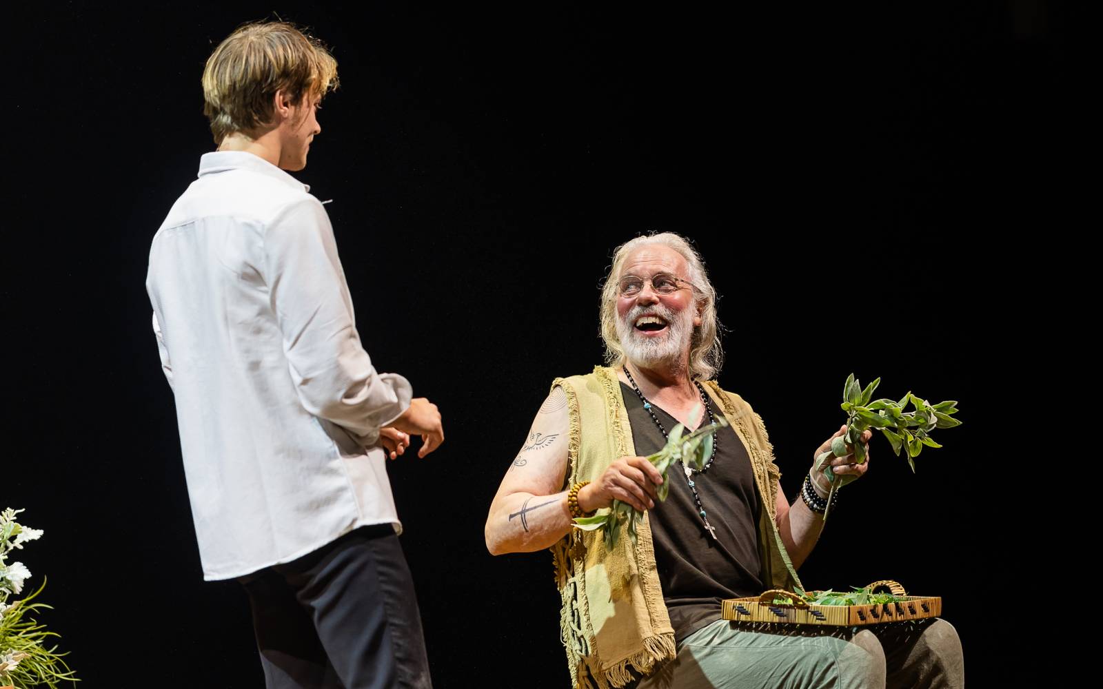 Rudy Pankow in a white dress shirt faces Terrence Mann, sitting and smiling broadly with herbs in his hand a small tray on his lap.