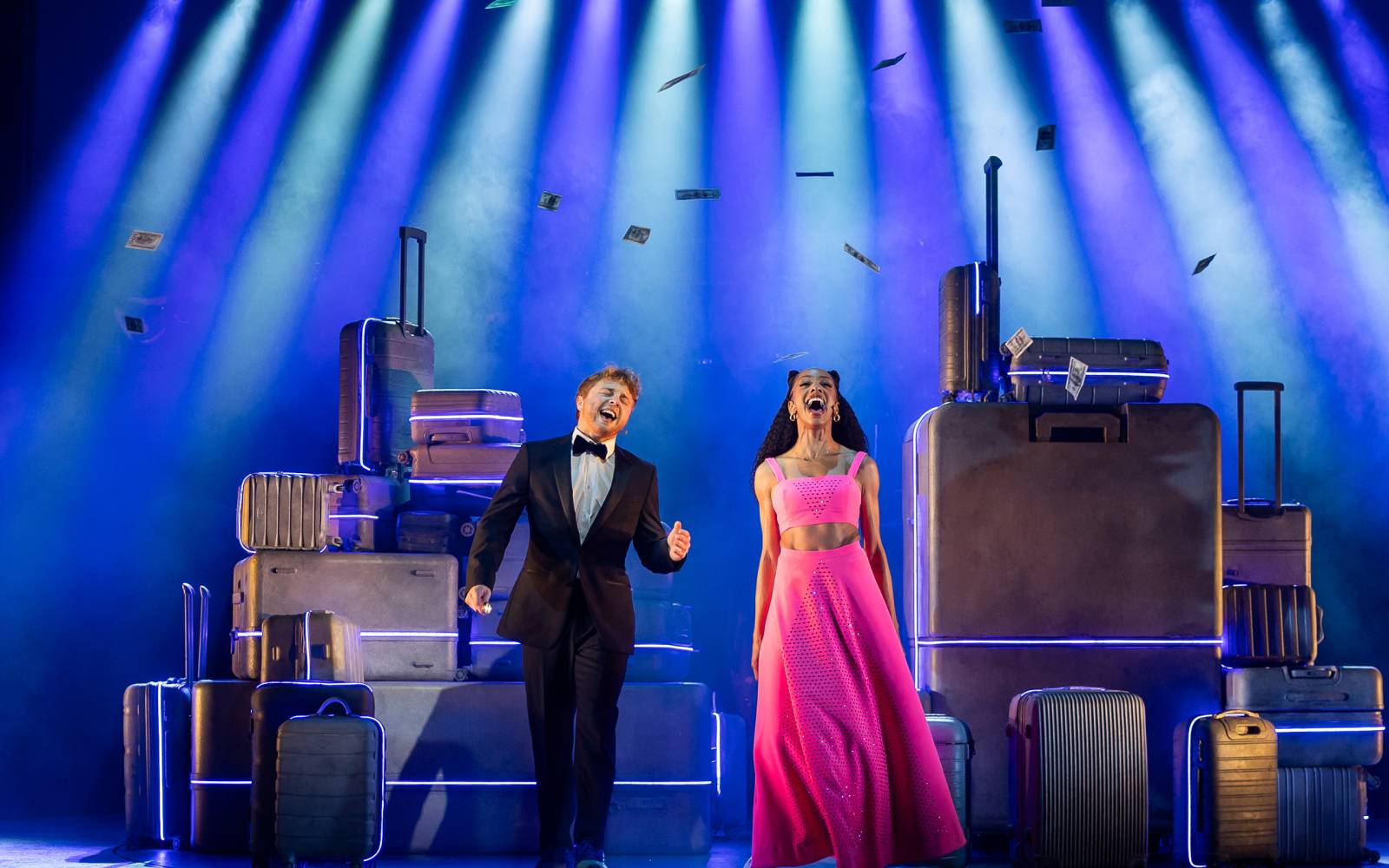Sam Tutty in a tuxedo and Christiani Pitts in a pink gown adorned with rhinestones stand center stage singing loudly under a wash of blue and white lights and dollar bills falling down on them.