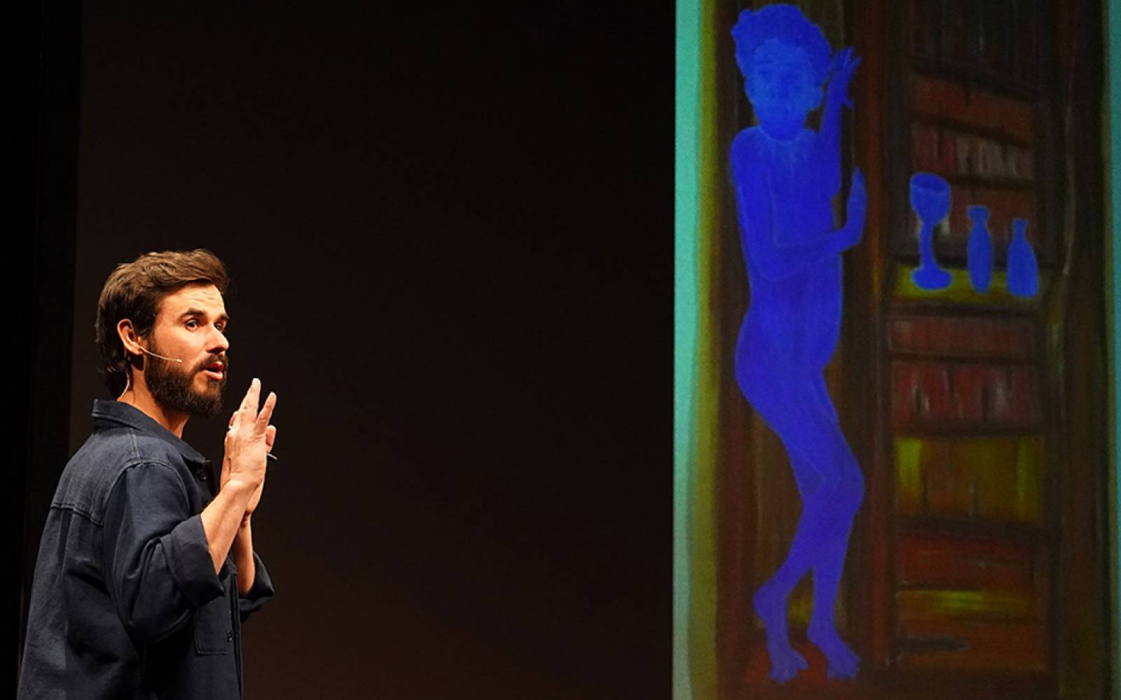 Sam Kissajukian in a dark grey flannel shirt raises his arms while speaking. To his right is a large paintings that features a bright blue human figure next to a black and green bookshelf.
