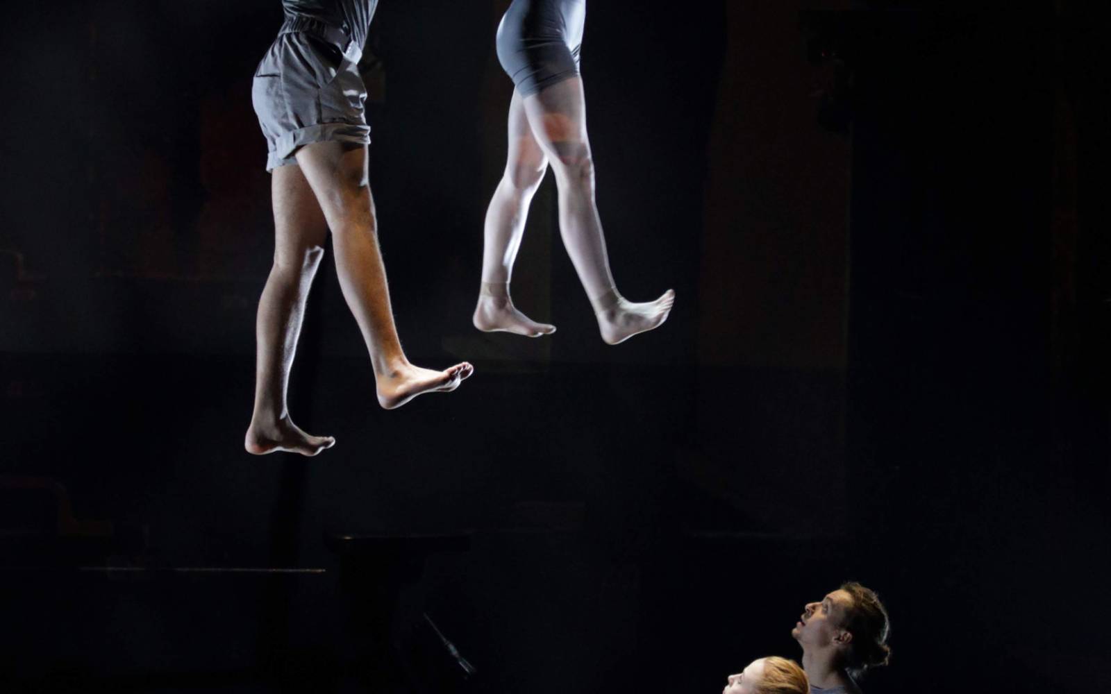 Two acrobats hang side-by-side from a trapeze by their hands with their feet flattened as if they're walking. Two performers on the ground look up at them.